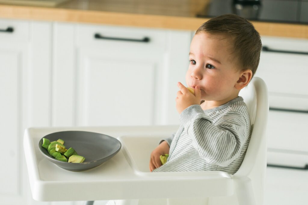 Happy baby sitting in high chair eating fruit in kitchen. Healthy nutrition for kids. Bio carrot as